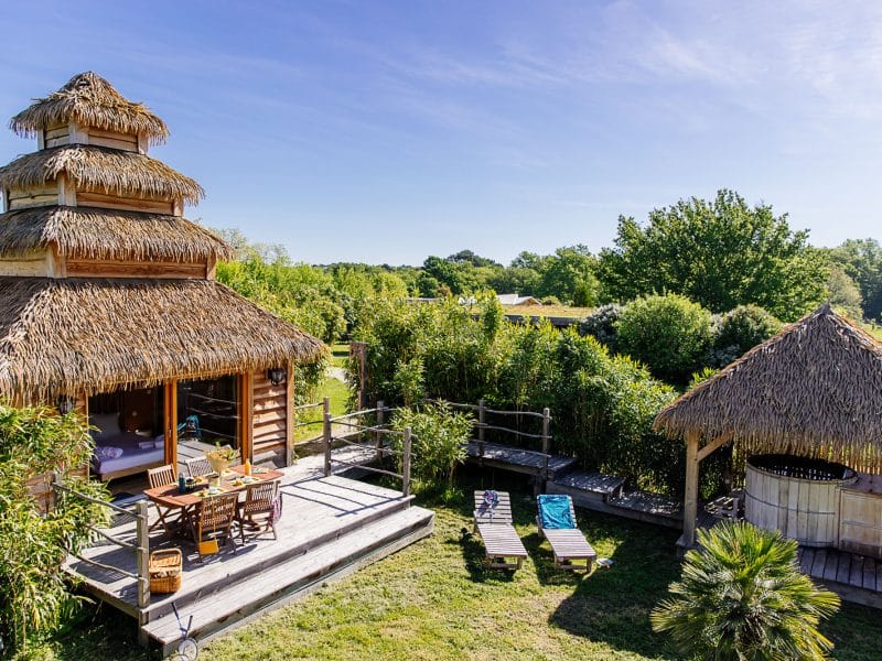 Cabane en bois avec toit de chaume, entourée de verdure et dun espace détente.