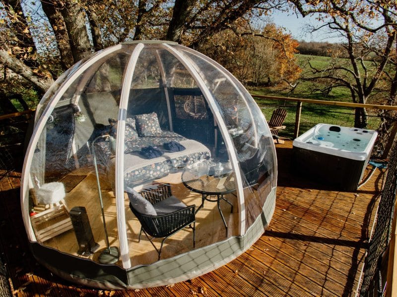 Dôme transparent sur une terrasse boisée, avec jacuzzi et vue sur la nature.