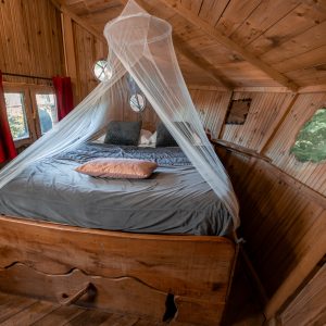 Cabane en bois avec lit douillet, entourée de fenêtres arrondies et dun voile léger.