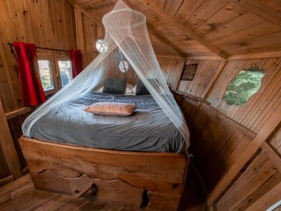 Cabane en bois avec lit douillet, entourée de fenêtres arrondies et dun voile léger.