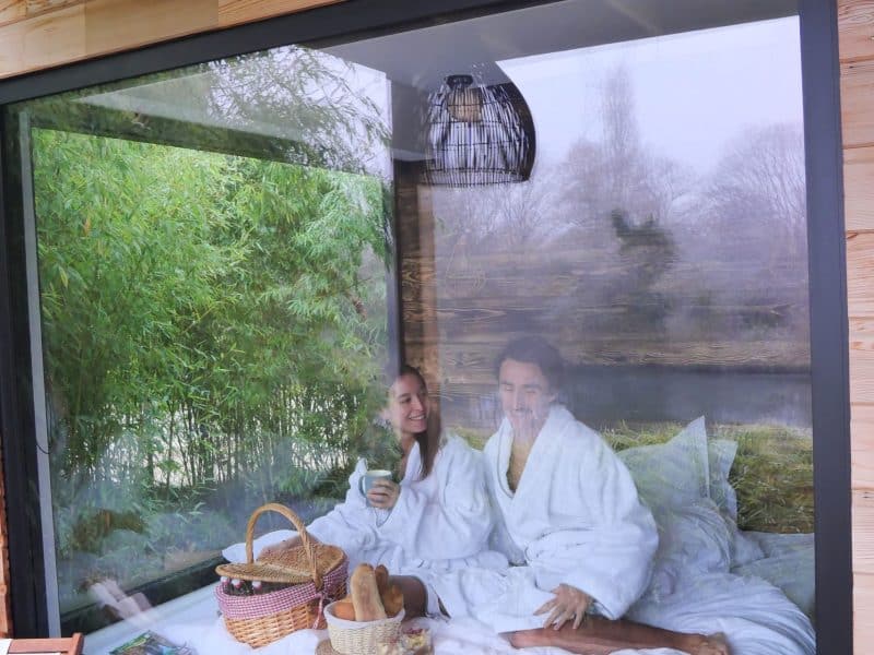 Cabane en bois avec grande fenêtre, couple en peignoir dégustant un petit-déjeuner.