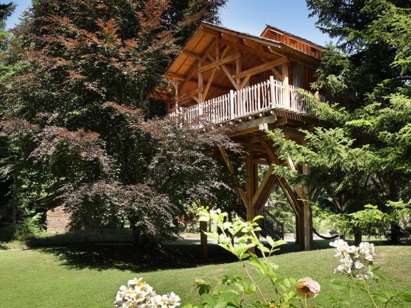 Cabane en bois perchée dans les arbres, entourée de verdure et de fleurs colorées.