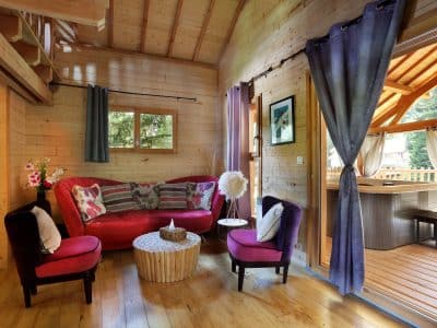Cabane en bois avec un salon cosy, canapé rouge et accès à une terrasse ensoleillée.