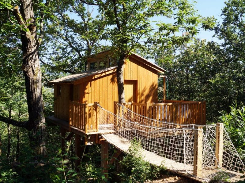 Cabane perchée en bois, entourée darbres, avec une passerelle en filet.