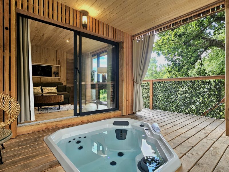 Cabane en bois avec jacuzzi sur la terrasse, entourée de verdure.