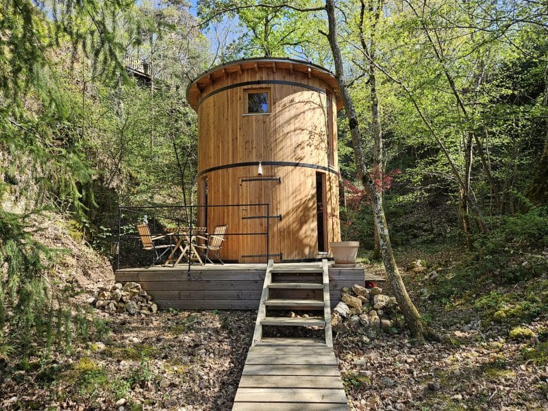 Cabane en bois ronde, entourée de verdure, avec terrasse et mobilier extérieur.