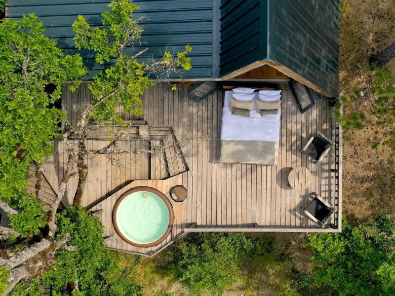 Cabane en bois avec terrasse en bois et jacuzzi, entourée darbres verdoyants.