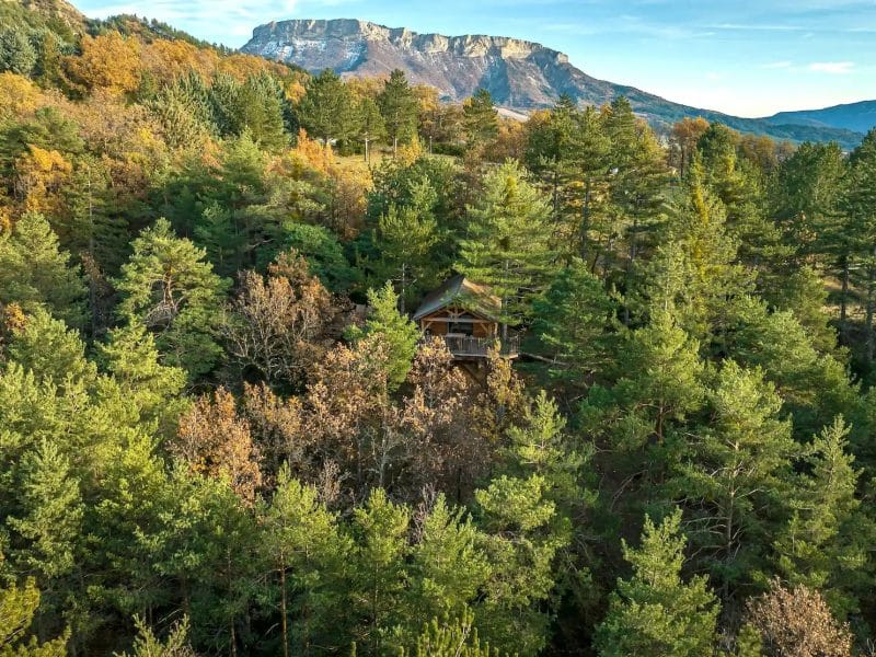 Cabane en bois nichée parmi les arbres, avec des montagnes en arrière-plan.