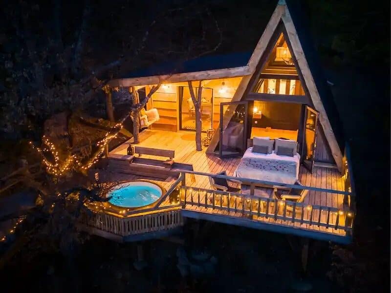 Chalet en bois avec jacuzzi extérieur, illuminé par des guirlandes dans la nuit.