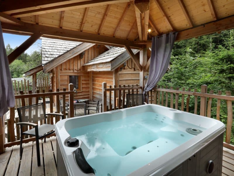 Chalet en bois avec jacuzzi sur la terrasse, entouré de verdure apaisante.
