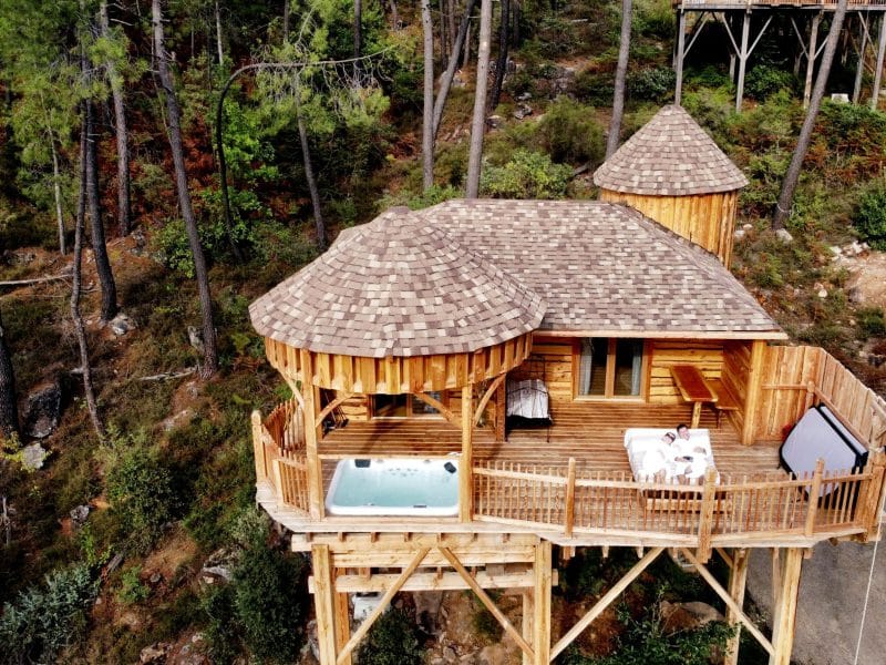Cabane perchée en bois avec jacuzzi, entourée de verdure et de pins.