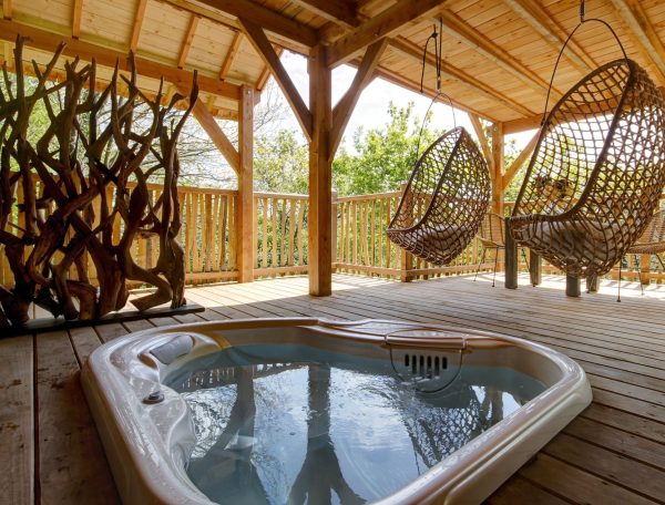 Cabane en bois avec jacuzzi et chaises suspendues, entourée de verdure.