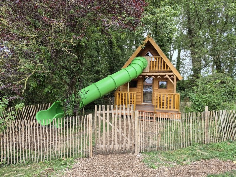 Cabane en bois avec toboggan vert, nichée dans la verdure des Hauts-de-France.