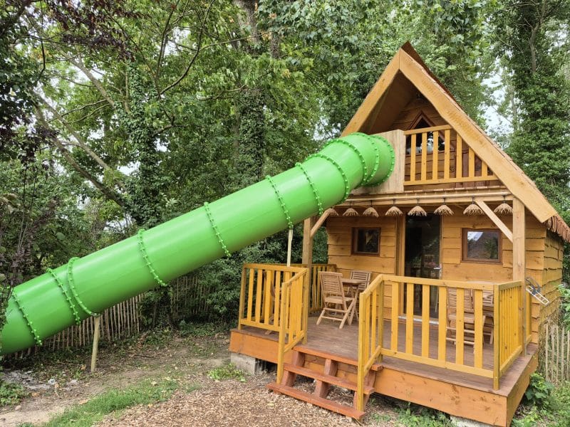 Cabane en bois avec toboggan vert, nichée dans la verdure. Parfait pour les familles.