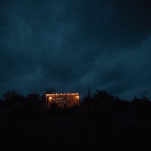 Cabane lumineuse dans la nuit, entourée darbres et de nuages sombres.