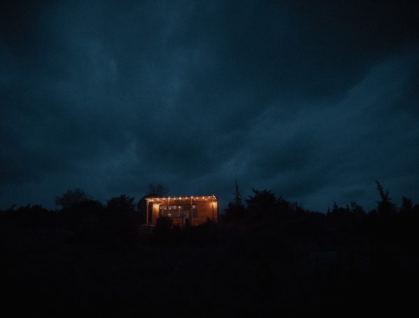 Cabane lumineuse dans la nuit, entourée darbres et de nuages sombres.