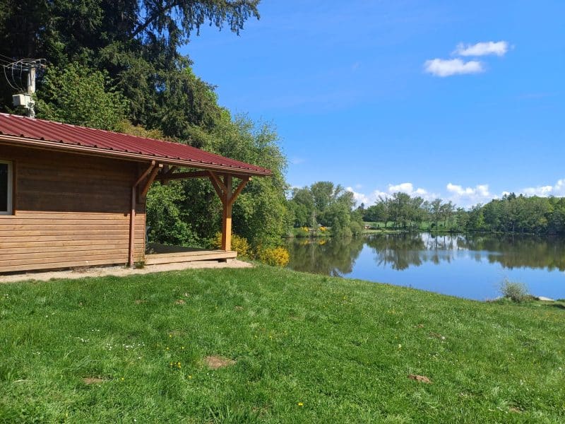 Cabane en bois au bord dun lac, entourée de verdure et sous un ciel bleu.