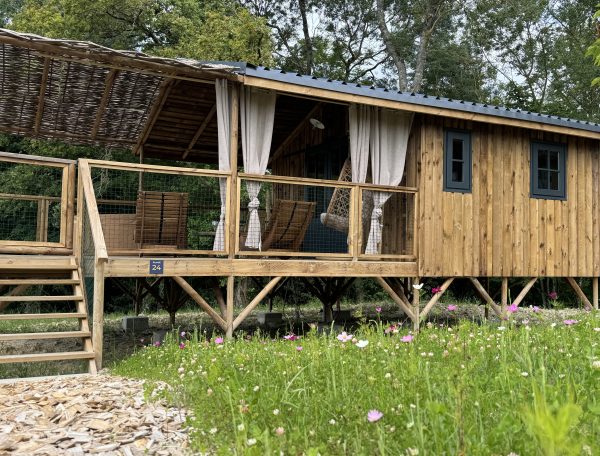 Cabane en bois sur pilotis, entourée de fleurs colorées et dune terrasse accueillante.