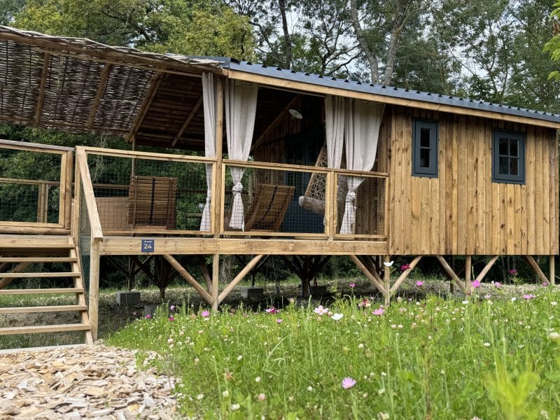 Cabane en bois sur pilotis, entourée de fleurs colorées et dune terrasse accueillante.