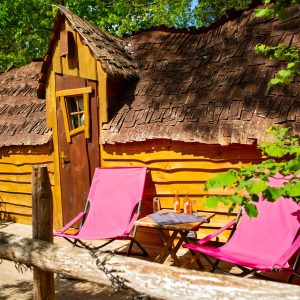 Cabane en bois avec toit en chaume, chaises roses et boissons sur la terrasse.