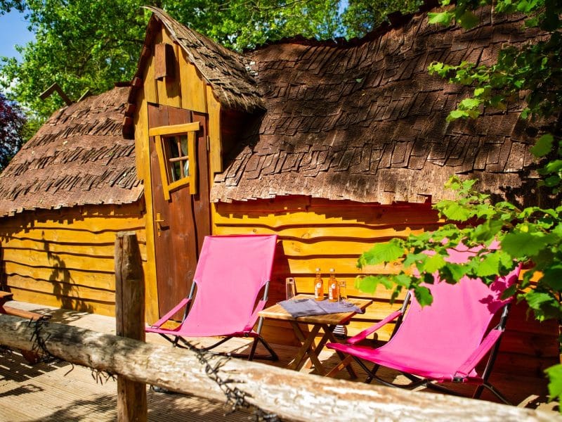 Cabane en bois avec toit en chaume, chaises roses et boissons sur la terrasse.