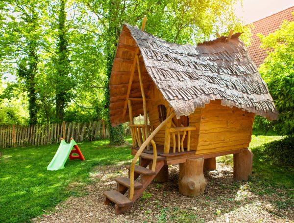 Cabane en bois dans un jardin verdoyant, avec toits en chaume et aire de jeux.