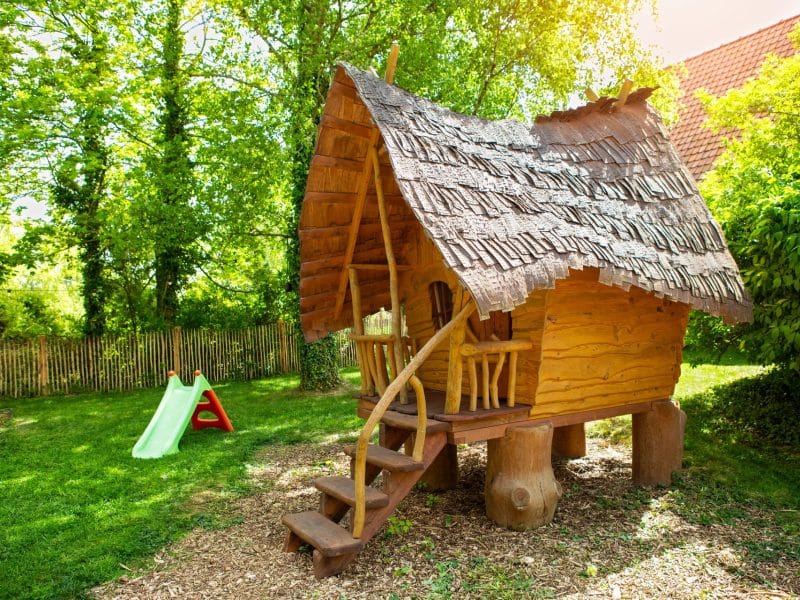 Cabane en bois dans un jardin verdoyant, avec toits en chaume et aire de jeux.