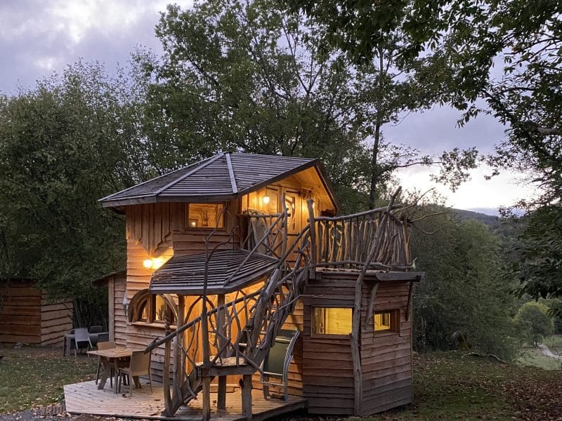 Cabane dans les arbres avec balcons et éclairage chaleureux, entourée de verdure.