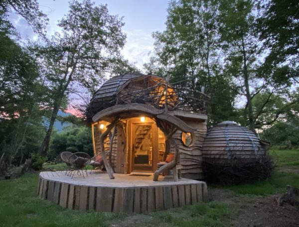 Cabane en bois unique, entourée darbres, avec une terrasse accueillante.