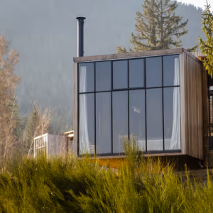 Cabane moderne avec grandes fenêtres, entourée de verdure et de montagnes.