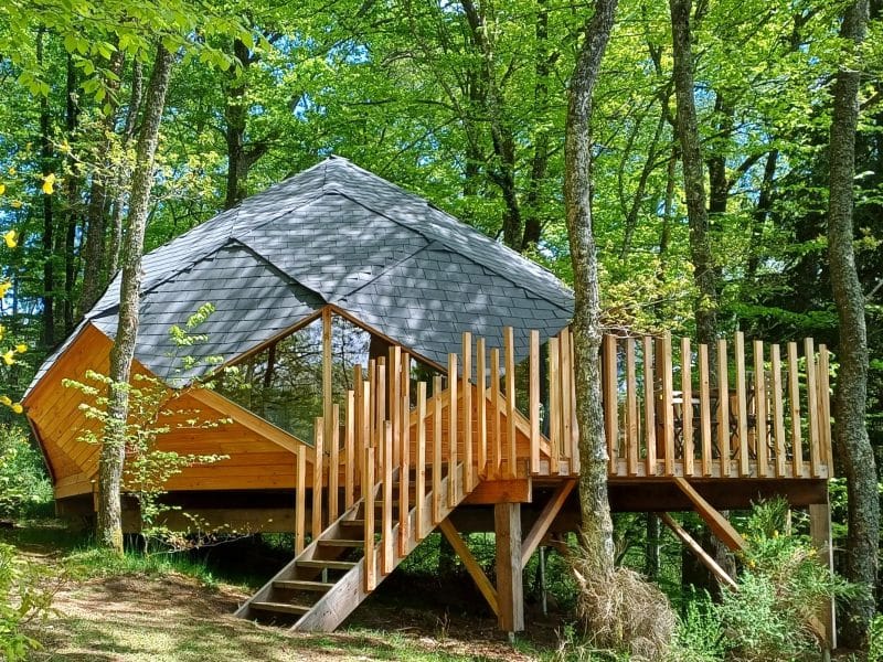Cabane en bois perchée, entourée darbres verdoyants, avec terrasse en bois.