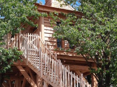 Cabane perchée en bois, entourée darbres, avec un accès par un escalier en bois.