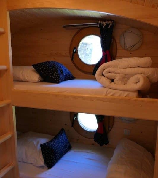 Chalet douillet avec lits superposés et fenêtres rondes, ambiance chaleureuse en bois.