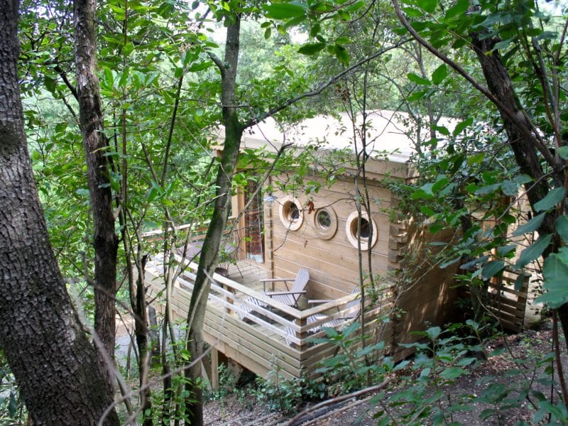 Cabane perchée en bois, entourée darbres, avec balustrade et fenêtres rondes.