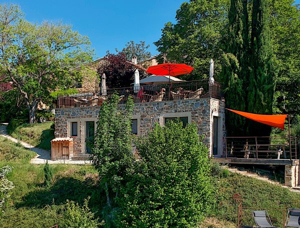 Maison en pierre avec terrasse ombragée et piscine, entourée de verdure.