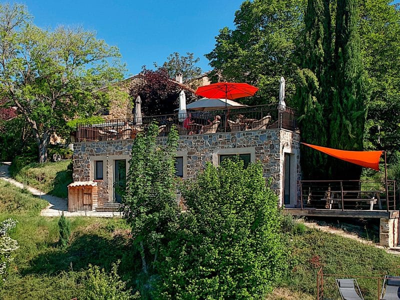 Maison en pierre avec terrasse ombragée et piscine, entourée de verdure.